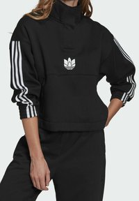 Svart bomullströja med hög krage, med vitt broderat logotyp och tre vita ränder på ärmarna. Strukturera cuffs och kortad design.