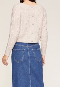 Beige gestrickter Pullover mit strukturierten Rautenmustern und dekorativen Knöpfen auf dem Rücken, kombiniert mit einem blauen Jeansrock.