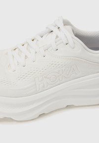 Vit träningssko med mesh-övre, texturerad logotyp och tjock sula. Har vita snören och subtila perforeringar för andningsförmåga.