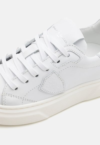 Baskets en cuir blanc dotées de lacets plats blancs, d'une texture lisse et d'un logo en relief subtil sur le côté ; semelle en caoutchouc avec un design texturé.