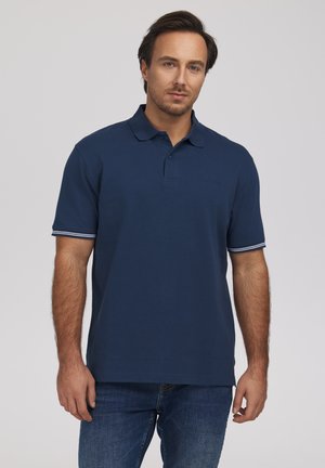 Mężczyzna z krótkimi ciemnymi włosami, ubrany w granatową koszulkę polo i niebieskie dżinsy, stojący na jednolitym jasnym tle.