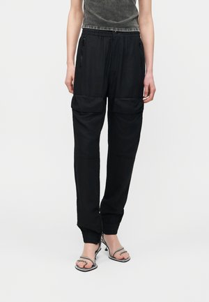 Άτομο που φοράει χαλαρό μαύρο cargo παντελόνι με τσεπάκια με φερμουάρ και μανσέτες στους αστραγάλους, σε συνδυασμό με μαύρα σανδάλια με τακούνι και λουράκια.