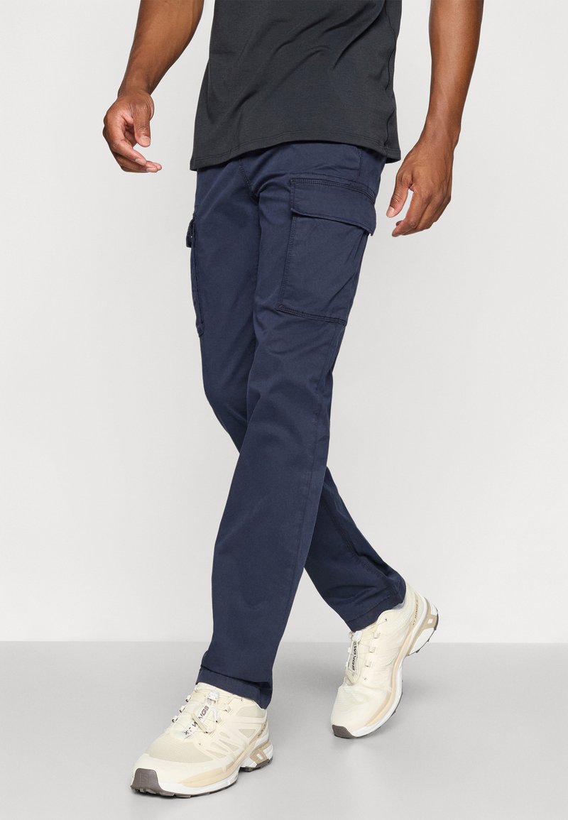 Un pantalon cargo marine en tissu robuste, doté de plusieurs poches, d'une coupe droite et d'une silhouette décontractée, associé à des baskets de couleur claire.
