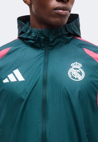 Gros plan d'une personne portant une veste zippée bleu sarcelle avec capuche, avec les logos blancs Adidas et Real Madrid et des touches roses sur les épaules.