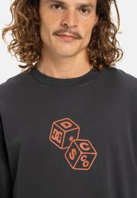 Černá košile s dlouhým rukávem vyrobená z bavlny, s oranžovou grafikou dvou kostek, na kterých jsou viditelné písmena 'D' a 'S'. Měkká textura.