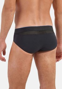 Schwarze Schwimmslips aus glattem Stoff, mit elastischem Bund und Netzakzenten sowie einer enganliegenden Passform, die die Körperkonturen des Trägers betont.