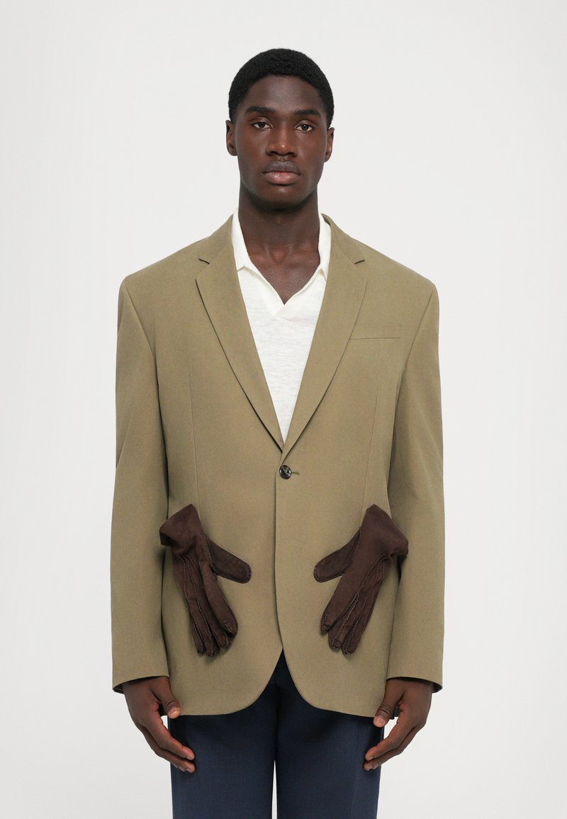 Olivgrüne Blazer mit braunen Lederhandschuhen, die am Bund befestigt sind. Darunter ein weißes Hemd, blaue Hosen und ein Ein-Knopf-Verschluss.