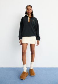 Φλις πουλόβερ σε σκούρο γκρι με καφέ γιακά και κουμπιά; συνδυασμένο με μια κοντή κρεμ φούστα και καφέ γούνινες παντόφλες.