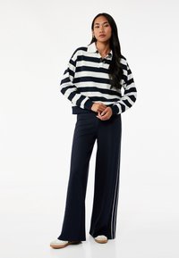Mørkeblå og hvidstribet pullover med krave og knap, parret med mørkeblå bukser med vide ben, der har hvide sidelommer og afslappet fodtøj.