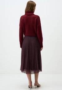 Maglione a collo alto borgogna con una vestibilità rilassata abbinato a una gonna plissettata con un motivo geometrico in blu navy e rosso.