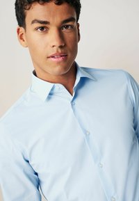 Chemise bleu clair à boutonnage avec un col ouvert, en tissu lisse, et boutons nacrés. L'arrière-plan est neutre et discret.