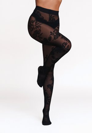 Jambes croisées portant des collants noirs transparents à motifs floraux, debout sur un pied sur un fond clair uni.
