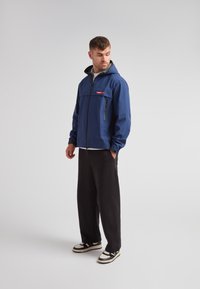 Veste à capuche bleue avec une fermeture éclair à l'avant, une poche sur la poitrine et une doublure grise. Associée à un pantalon noir et des baskets avec des accents noirs et blancs.