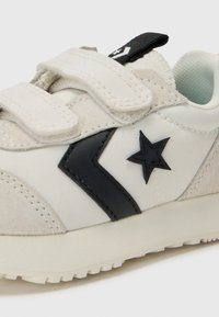 Baskets pour enfants avec un design blanc et beige, présentant des accents noirs, deux bandes velcro et un logo en forme d'étoile sur le côté.