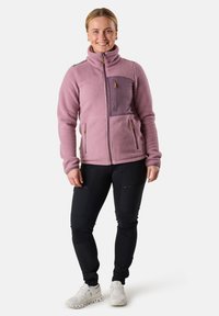 Fleece jacket in lichtroze met een hoge kraag, voorzien van een donkerpaarse zak en twee zijzakken. Gemaakt met zwarte broek en witte schoenen.