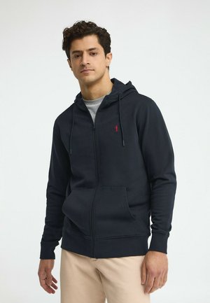 Hombre vistiendo una sudadera con capucha azul marino con cremallera y bolsillos delanteros, y pantalones beige, de pie frente a un fondo claro y sencillo.