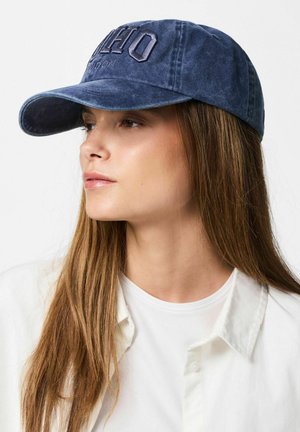 Kvinne med langt brunt hår som har på seg en blå baseballcaps med brodert "OHIO London", hvit skjorte, ser mot venstre.