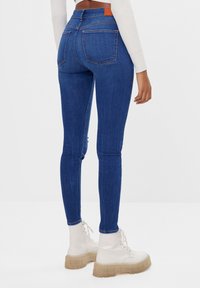 Jeans ajustados de mezclilla azul oscuro con cintura alta. Presenta dos bolsillos traseros y un detalle de cuero marrón en la cinturilla.