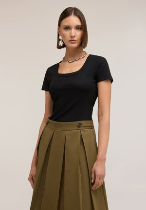 Frau mit glatten braunen Haaren trägt ein schwarzes kurzärmeliges geripptes Oberteil, einen olivgrünen plissierten Rock mit Knopfdetails und silbernen Schmuck.