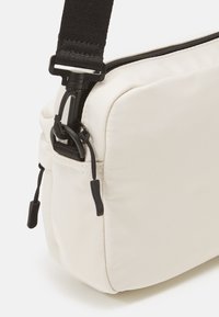 Hvid stof crossbody taske med sort rem, udstyret med lynlås, struktureret overflade og justerbar spænde. Rundet form og enkelt design.