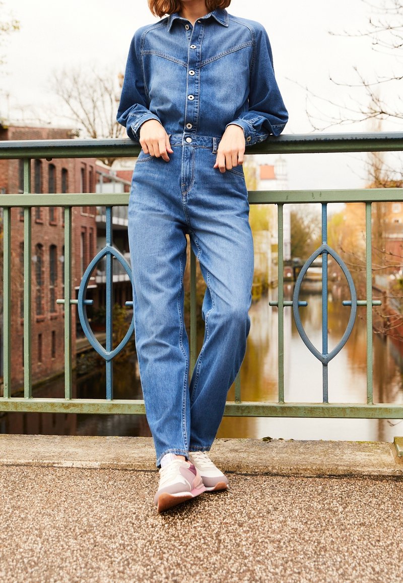 Personne en chemise en jean bleue et en jeans debout sur un pont, appuyée à la rambarde, les bras croisés, avec une rivière en arrière-plan.
