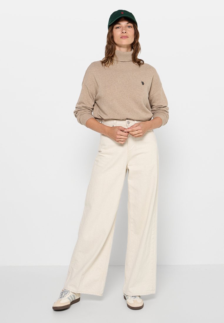 Maglione a collo alto beige, pantaloni larghi crema e berretto verde. L'outfit presenta un design simple, una texture morbida e un branding discreto.