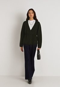 Tmavě zelená pletená kardigánová bunda s vázáním v pase, spárovaná s bílou košilí a tmavými kalhotami s širokými nohavicemi; drží černou kabelku zdobenou cvočky.