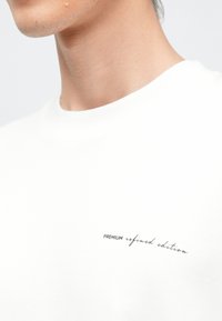 Λευκό βαμβακερό t-shirt με στρογγυλή λαιμόκοψη και διακριτικό κείμενο στο στήθος που γράφει «PREMIUM refined edition» σε καλλιγραφική γραμματοσειρά.