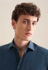 Korte mouw, donker turquoise shirt met een knoopsluiting aan de voorkant, subtiele bloemenpatroon bij de kraag, gladde stof en een enkele borstzak.