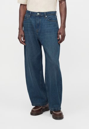 Personne portant un jean bleu ample, des chaussures marron style mocassin et une chemise beige clair à manches courtes sur un fond uni.