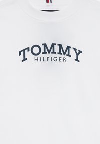 Hvid skjorte med "TOMMY HILFIGER" trykt i sort, med rund halsudskæring og et lille rødt, hvidt og sort stribet mærke indeni kraven.