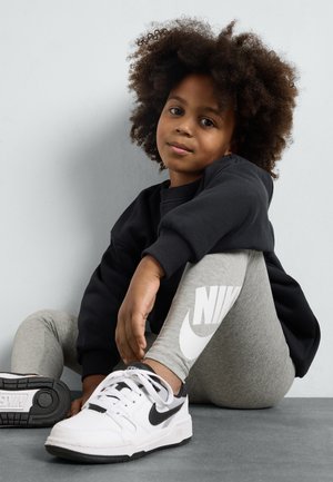 Svart hettegenser, grå leggings med Nike-logo, og hvite joggesko med svarte detaljer. Strukturert krøllete hår. Sitter mot en lys bakgrunn.