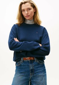 Frau mit blonden Haaren, die einen marineblauen Pullover über einem grauen Rollkragenpullover trägt, blaue Jeans und einen braunen Gürtel mit einer Metallschnalle hat, die Arme verschränkt.