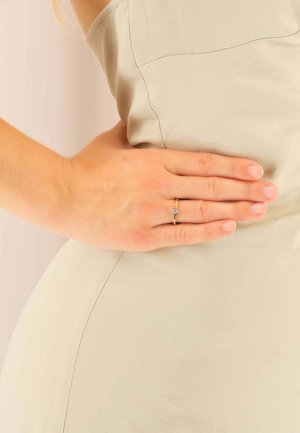 Bague en or avec une pierre transparente, portée sur un doigt, avec une peau lisse et un fond en tissu beige. Le tissu semble léger et texturé.