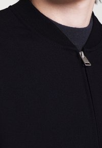 Giubbotto bomber nero con colletto a costine, chiusura con zip metallica e texture liscia, che mette in risalto un design moderno e minimal.