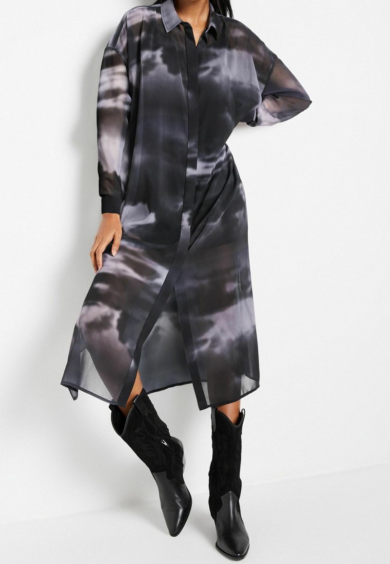 Femme portant une longue robe tie-dye noire et grise à manches longues et des bottes de cowboy noires à bout pointu, debout sur un fond blanc.