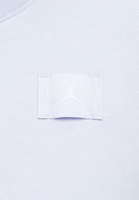 Világoskék textúrált anyag, amelyen egy fehér, hímzett Jordan logó címke található, középen a ruhadarabon.