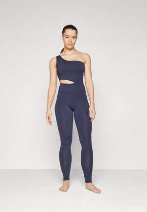 Top de activewear asimétrico en color marino con un detalle recortado, combinado con leggings de talle alto y ajustados en tejido de similar tonalidad oscura.