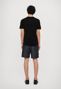 Man with curly hair wearing a black short-sleeve shirt, dark rolled-up shorts, and black shoes, standing facing a white background.