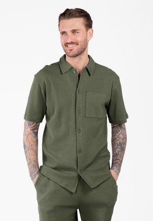 Man met getatoeëerde armen in een olijfgroene kortegelijfde overhemd met bijpassende broek, handen in de zakken, licht glimlachend, op een witte achtergrond.