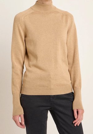 Pull à col roulé beige avec manches longues et poignets côtelés porté avec un pantalon noir, raccourci au niveau du milieu du torse.
