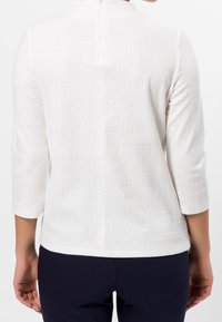 Top bianco con texture e maniche a tre quarti, dotato di zip centrale sul retro e scollatura rotonda.