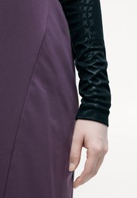 Garums piedurkņu tops ar teksturētu melnu audumu un gludu purpura kleitas daļu, kurai ir izteikta šuve jostasvietā.