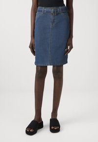 Denim kokerrok in blauw, met een tailleband, zakken aan de voorkant en een knielange snit. Draagt zwarte slides.