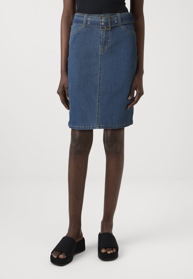 Denim kokerrok in blauw, met een tailleband, zakken aan de voorkant en een knielange snit. Draagt zwarte slides.