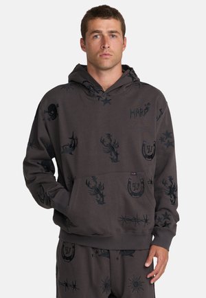 Man die een donkergrijze hoodie en bijpassende broek draagt met zwarte tattoo-achtige ontwerpen waaronder sterren, schedels, schorpioenen en hoefijzers.
