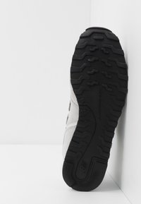 Chaussure de sport grise en daim avec une semelle extérieure en caoutchouc noir présentant un motif texturé et une marque au bas. Vue latérale claire de la semelle.