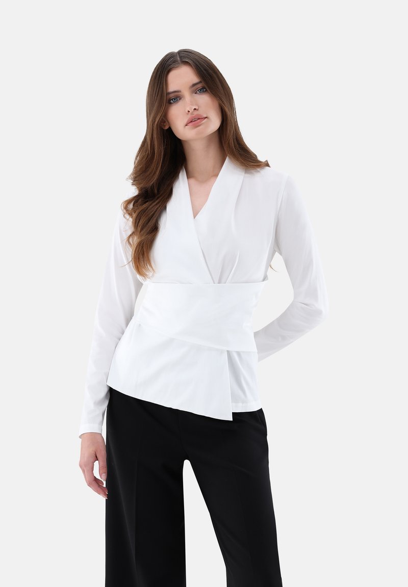 Blusa avvolgente bianca con maniche lunghe, scollatura strutturata e vita a peplum. Abbinata a pantaloni neri per contrasto.