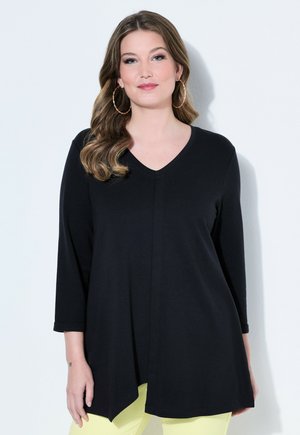 Frau mit welligen braunen Haaren und Creolen trägt eine schwarze V-Ausschnitt-Tunika mit dreiviertellangen Ärmeln und blassgelbe Hosen, steht vor einer weißen Wand.