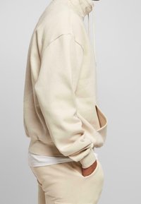 Persona che indossa un hoodie beige e pantaloni abbinati con le mani in tasca, una camicia bianca visibile sotto.